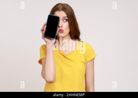 Look, advertise here. Portrait of funny woman of young age in T-shirt covering half of face phone with blank screen,has thoughtful doubting expression. Indoor studio shot isolated on gray background. Stock Photo