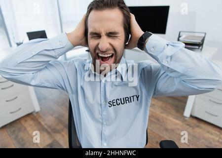 shocked security man with closed eyes holding hands on head and shouting in office Stock Photo