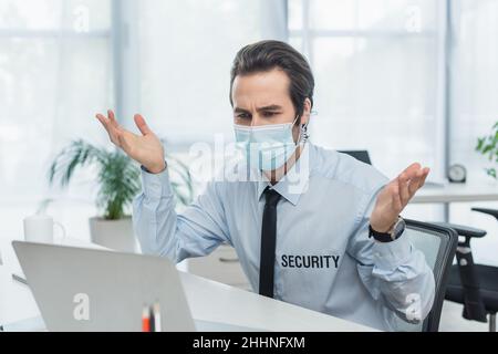 discouraged security man in medical mask gesturing near blurred laptop Stock Photo