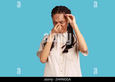 Portrait of frustrated woman with black dreadlocks touching closed eyes, crying from depression, grief or pain, feeling hopeless, wearing white shirt. Indoor studio shot isolated on blue background. Stock Photo