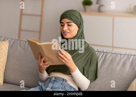 Peaceful domestic hobbies. Cheery young Arab woman in hijab reading book, relaxing on sofa in living room Stock Photo