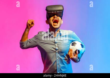 Portrait of man football fan cheering up supporting favorite team with soccer ball, watching match in vr headset, doing winner gesture. Indoor studio shot isolated on colorful neon light background. Stock Photo