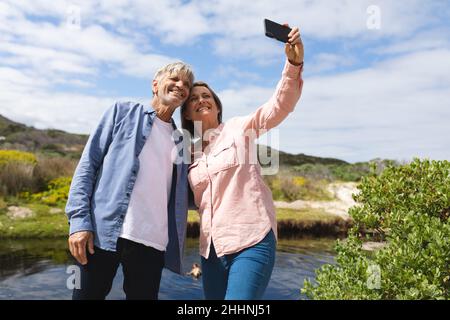 Cheerful caucasian couple taking selfie through smart phone at lakeshore against sky during weekend. togetherness, weekend, wireless technology and na Stock Photo