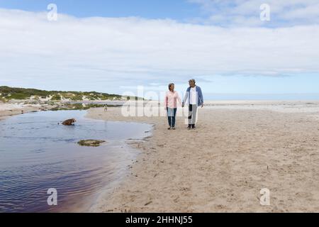 Full length of caucasian mature couple holding hands while walking on sand at beach during weekend. togetherness, weekend and nature concept. Stock Photo