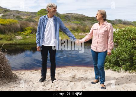 Happy caucasian mature couple holding hands and looking at each other while standing at lakeshore. togetherness, weekend and nature concept. Stock Photo