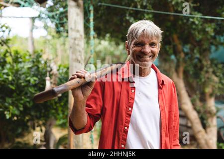 Portrait of happy caucasian mature man carrying shovel while standing in backyard. lifestyle and farming concept. Stock Photo