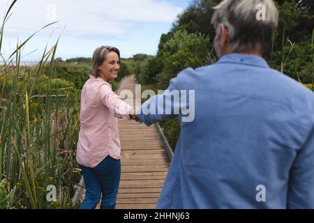 Happy caucasian mature couple holding hands while standing on boardwalk during weekend. togetherness, weekend and nature concept. Stock Photo