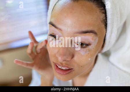 Closeup of biracial young woman in bathrobe applying cream on face. unaltered, routine, lifestyle, skin condition and vitiligo. Stock Photo