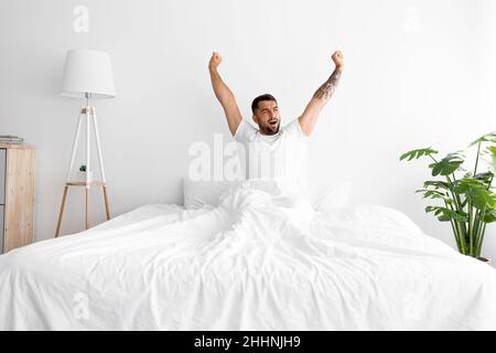 Satisfied smiling relaxed young caucasian muscular man sitting on white bed, stretching body in minimalist bedroom Stock Photo