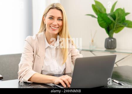 Smiling business woman using laptop for remote work from home. Confident lady checking emails, conducting business correspondence while sitting at the table, looks at the camera Stock Photo