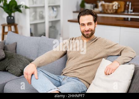 Portrait of handsome young man having rest in modern living room at home, sitting casually on sofa, looking at the camera, feeling relaxed. Lifestyle and leisure concept Stock Photo