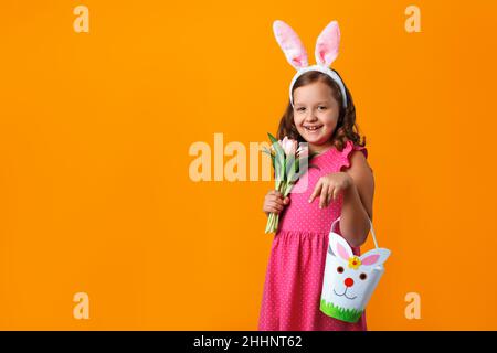 A cute little girl in Easter bunny ears holds a bag for sweets and a bouquet of tulips. Portrait of a child on a yellow isolated background. Copy spac Stock Photo