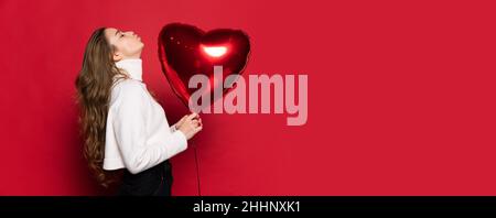 Young pretty with long wavy silky hair, natural makeup holding red heart balloon looking up isolated on red background. Valentines Day concept Stock Photo