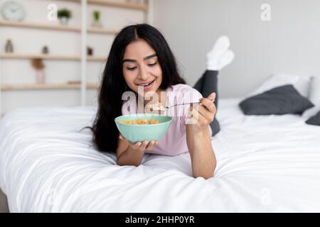 Portrait of slim young Indian lady eating cereal with milk while lying on bed at home, full length Stock Photo
