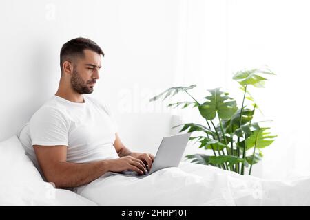 Busy serious concentrated young caucasian muscular businessman sit on white bed working on computer Stock Photo