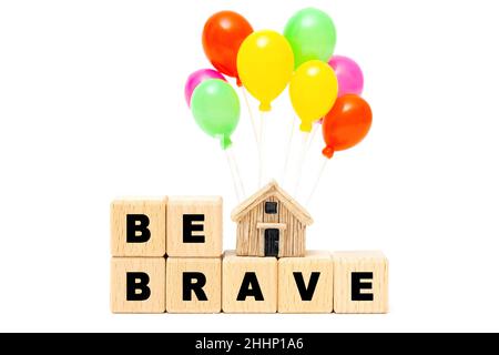 Text BE BRAVE made from wooden letter blocks and a miniature house model with toy party balloons isolated on white background. Overcoming the fear of Stock Photo