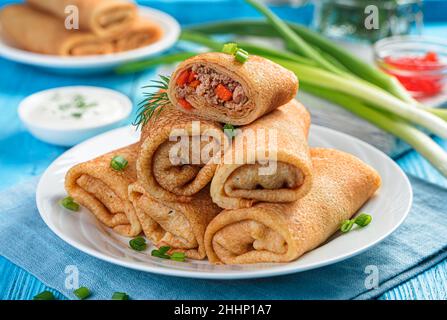 Thin fried pancakes stuffed with minced meat with carrots and decorated with fresh dill and onions on a blue background. Stock Photo