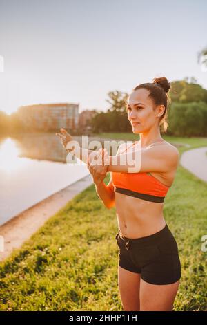 Fitness woman stretching arms, warming up near lake on sunny summer morning Stock Photo