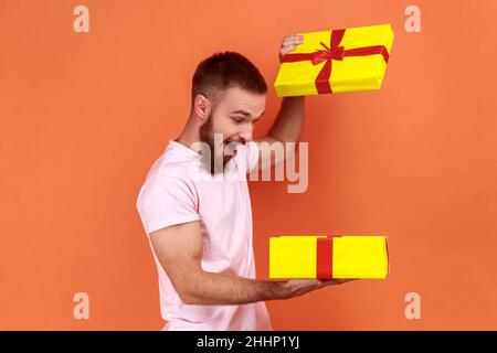 Portrait of handsome bearded man holding in hands unboxed gift, being satisfied with present, being extremely happy, wearing pink T-shirt. Indoor studio shot isolated on orange background. Stock Photo