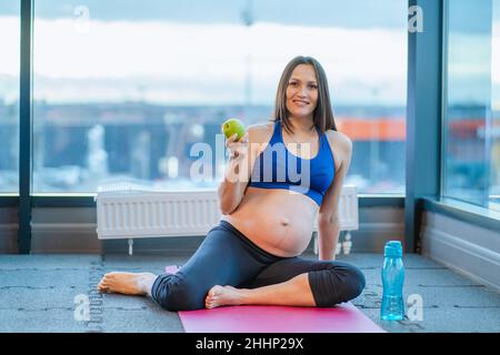 Happy pregnant woman sitting on yoga mat with apple while exercising at gym Stock Photo