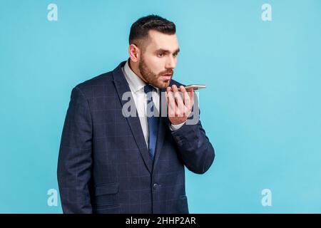 Serious bearded businessman wearing official style suit holding mobile phone in hand, giving commands to voice assistant, leaving voice message. Indoor studio shot isolated on blue background. Stock Photo