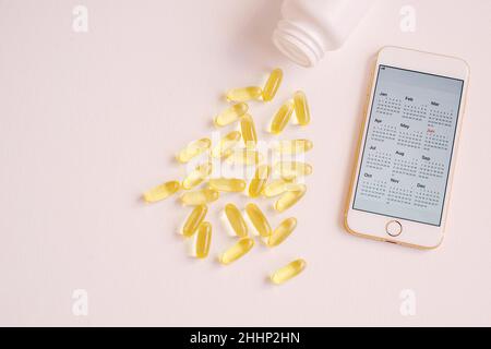 A lot of pills are spilled of a plastic container and a smartphone with calendar screen. Close up, top view with pink neutral background. Vitamin ther Stock Photo
