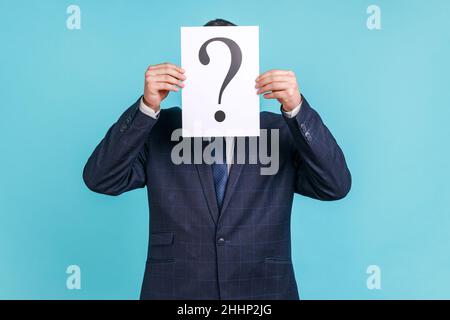 Portrait of anonymous male wearing dark official style suit covering his face with paper with question mark, finding right solution. Indoor studio shot isolated on blue background. Stock Photo