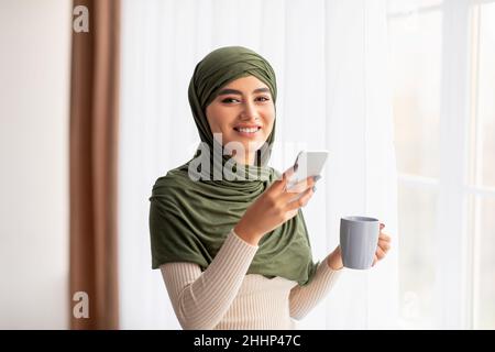 Smiling young Arab female in hijab standing near window with coffee and cellphone, checking social network, browsing web Stock Photo