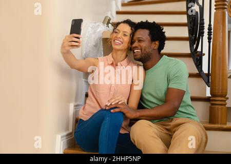 Cheerful young multiracial couple sitting on staircase while taking selfie at new house. people, relocation and technology concept, unaltered. Stock Photo