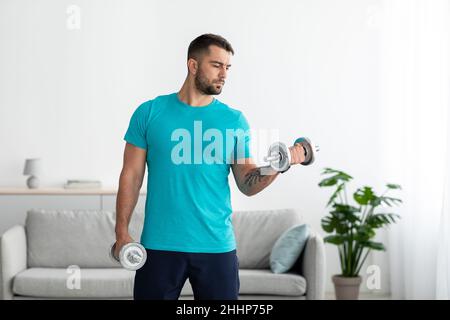 Concentrated young caucasian man athlete in blue t-shirt lifts dumbbells and does exercises for biceps Stock Photo