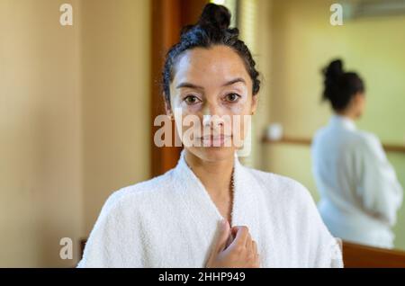 Portrait of biracial young woman wearing bathrobe standing in bathroom. unaltered, routine, lifestyle and vitiligo. Stock Photo