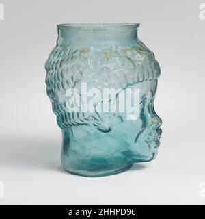 Glass cup in the form of the head of a Black African 2nd half of 1st century A.D. Roman Translucent blue green.Lopsided, everted, thick, knocked-off rim; narrow, cylindrical neck; body modeled in the shape of a head in the round; base ring with rounded lower edge; flat bottom. Two mold seams run from rim down sides of head to base ring, with a separate disk-shaped base section.On body, a head wearing an ivy wreath and globular pendant earrings; one half of mold comprises the back of the head and part of the proper left ear, with the hair arranged in six horizontal rows of short vertical locks Stock Photo