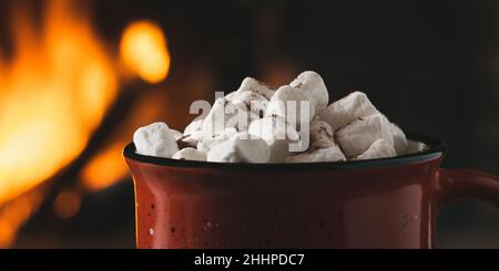 Cocoa with marshmallows and chocolate in a red mug near a burning fireplace. Stock Photo