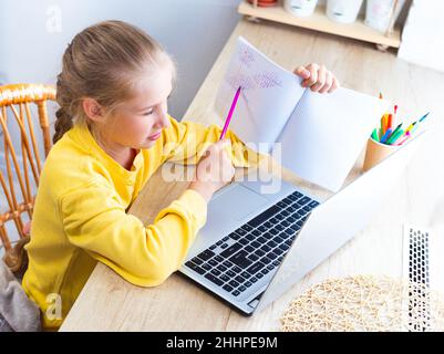 Caucasian cute schoolgirl in yellow sweatshirt doing her math homework at home in front of laptop, showing the solved example to teacher on screen. E Stock Photo