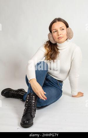 Sitting attractive young european woman with brown hair in warm white jumper isolated on white with fur earmuffs Stock Photo