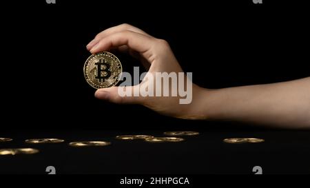 Woman's hand holds a bitcoin over a black table on which other coins lie. Stock Photo