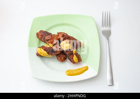 Mici or mitititei (traditional romanian food) with mustard on a ceramic plate, on white background. Hand holding a stainless steel fork Stock Photo