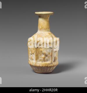 Glass hexagonal bottle ca. A.D. 25–50 Roman Opaque white.Rim folded out, round, and in, then pressed into flaring mouth; misshapen cylindrical neck; downward sloping shoulder; hexagonal body with vertical sides, then cup-shaped below; low base with rounded edge and slightly indented bottom. Mold seams run from base of neck, down sides, and meet at center of bottom.Decoration in relief in three registers: on shoulder, six downturned rays or pointed petals; on body, six rectangular panels, each decorated at top with a triangular pediment flanked by prominent but indistinct round objects, framed Stock Photo