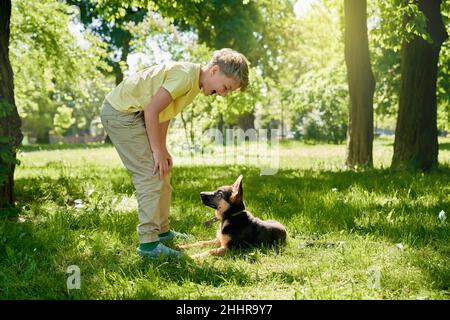 Cheerful little boy in casual wear playing with his little puppy at green city park. Caucasian child enjoying time spending with pet on fresh air. Sunny weather outdoors. Stock Photo