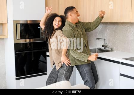 Curly woman and her guy dancing in the kitchen and singing. Merry multiracial couple having fun, fooling around while preparing dinner at home. Humor concept Stock Photo