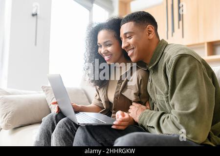 Young multiracial couple looking at the screen while using laptop on the sofa at home. Spouses watching something on laptop. Happy man and woman embracing and relaxing Stock Photo