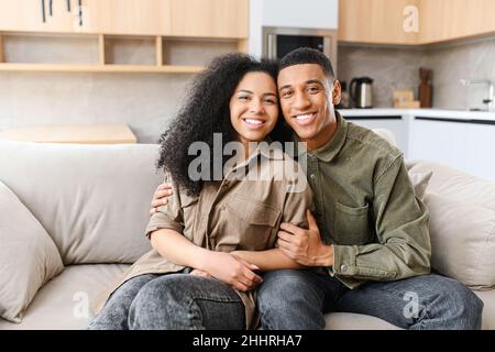 Happy young couple sitting on couch in living room and looking at camera at home. Smiling millennial man and woman having pleasure time together Stock Photo