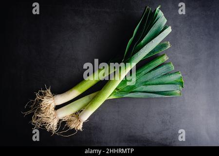 Overhead View of Whole Leeks on a Dark Background: Three leeks with leaves and roots on a stone countertop Stock Photo