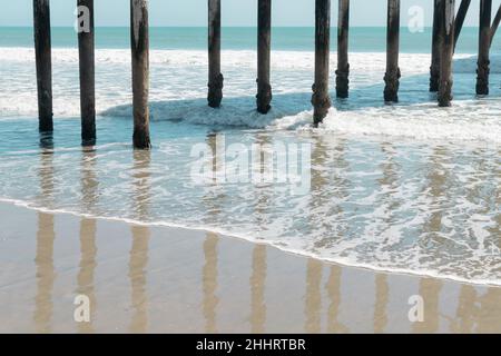 Pier pilings reflected on a wet, sandy beach and waves in San Simeon, California Stock Photo