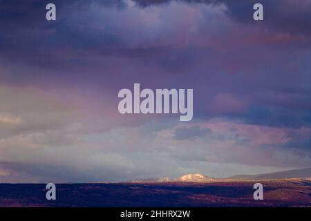 Dark purple storm clouds over a vast desert landscape with snow-capped mountains illuminated by sunset above a village in northern New Mexico Stock Photo