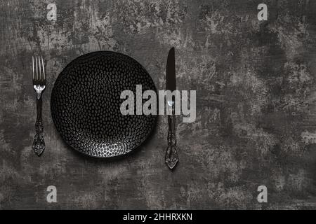 Set table. Black dinner plate, table knife and fork over dark textured surface. Empty porcelain plate near vintage knife and fork for food design. Stock Photo