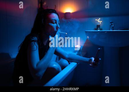 Young nicotine addicted woman with smartphone smoking cigarette, sitting in bathtub under neon light Stock Photo