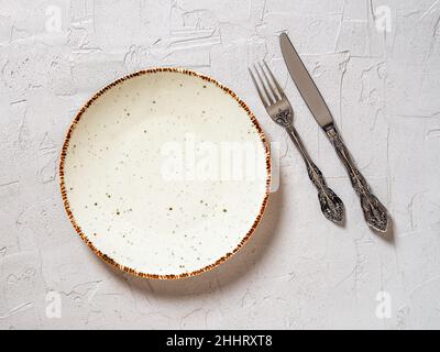 White porcelain dinner plate, vintage table knife and fork on a textured gray background. Empty speckled plate near elegant knife an fork. Top view. Stock Photo