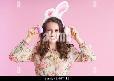 young woman with long wavy brunette hair with bunny ears and easter egg against pink background. Stock Photo