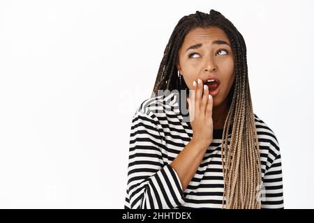 Close up portrait of black girl yawning, looking aside with unamused bored face expression, standing in striped blouse over white background Stock Photo